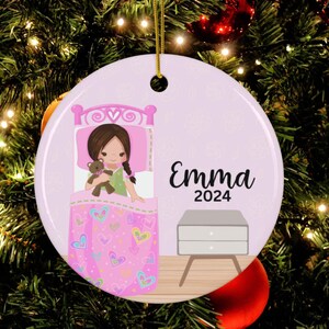 May include: A pink and white personalized Christmas ornament featuring a girl in bed with a teddy bear. The ornament has the name "Emma" and the year "2024" printed on it.