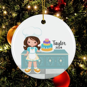 May include: Round white Christmas ornament featuring a cartoon girl in a chef's hat and apron holding a tiered cake. The ornament has the name "Taylor" and the year "2024" printed on it. The background is a Christmas tree.