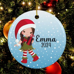 May include: A blue and white Christmas ornament with a cartoon elf girl holding a gift. The elf is wearing a red hat, green dress, and red and white striped leggings. The ornament has the name "Emma" and the year "2024" written on it.