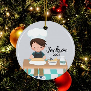 May include: Round ceramic Christmas ornament featuring a cartoon chef rolling dough. The ornament has the name "Jackson" and the year "2024" printed on it. The ornament is hanging from a gold string.