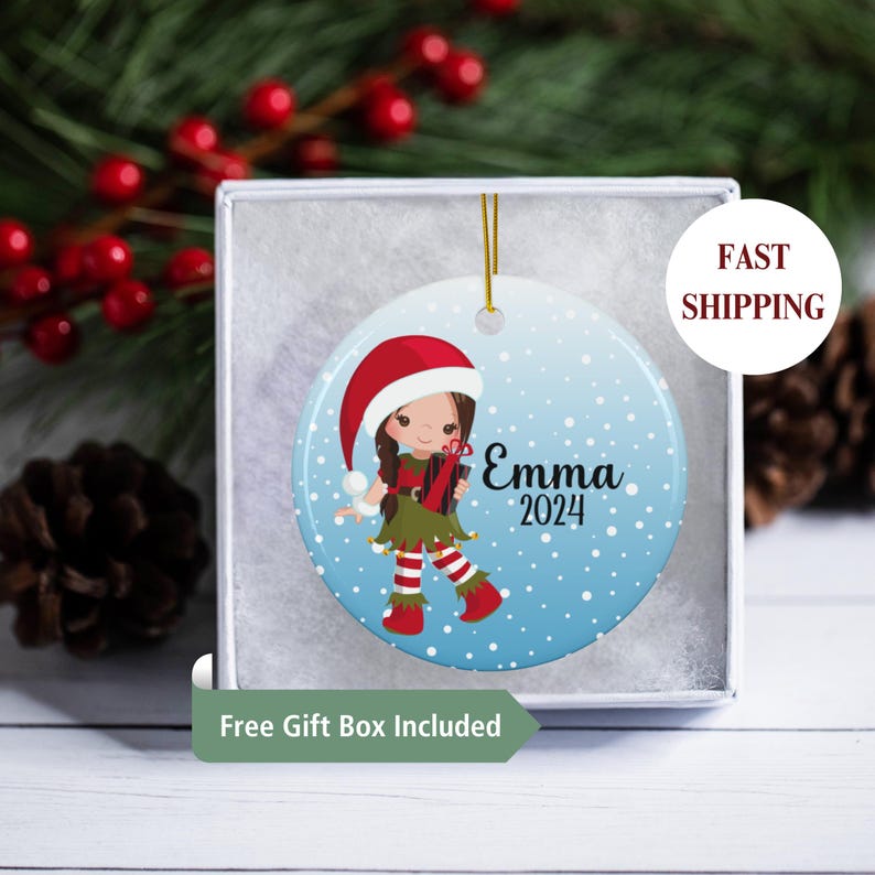 May include: A blue and white Christmas ornament with a cartoon elf girl holding a gift. The ornament has a red Santa hat, brown hair, and the name "Emma 2024" written on it.