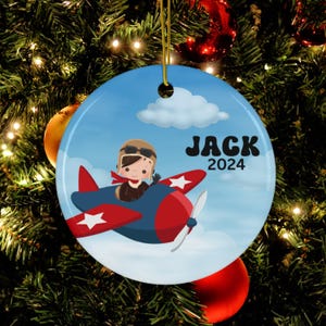 May include: A blue and red airplane ornament with a white star on the wing. A cartoon pilot with brown hair is in the cockpit. The ornament has the name "JACK" and the year "2024" printed on it.