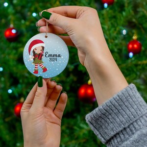 May include: A round blue and white Christmas ornament with a cartoon girl wearing a red Santa hat and green and red elf outfit. The ornament has the name "Emma" and the year "2024" printed on it.