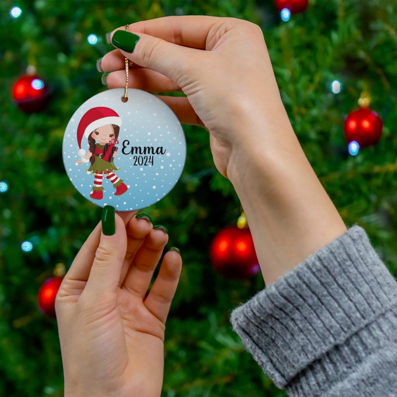 May include: A round blue and white Christmas ornament with a cartoon girl wearing a red Santa hat and green and red elf outfit. The ornament has the name "Emma" and the year "2024" printed on it.