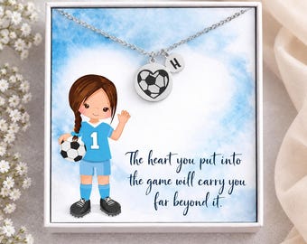 Könnte beinhalten: Eine silberfarbene Halskette mit einem herzförmigen Fußball-Anhänger und einem runden Anhänger mit dem Buchstaben H. Der Schmuck liegt in einer weißen Schachtel mit einer Illustration einer Fußballspielerin und dem Text: The heart you put into the game will carry you far beyond it.