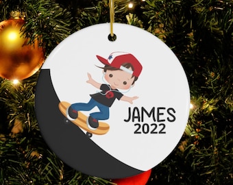 May include: A white ceramic Christmas ornament with a cartoon illustration of a boy skateboarding. The boy is wearing a red hat, black shirt, blue jeans, and headphones. The ornament has the name "JAMES" and the year "2022" printed on it.