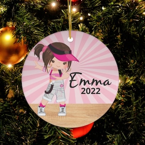 May include: A pink and white Christmas ornament featuring a cartoon girl wearing a pink hat and overalls. The ornament has a pink and white striped background with a woodgrain base. The text "Emma 2022" is printed on the ornament.