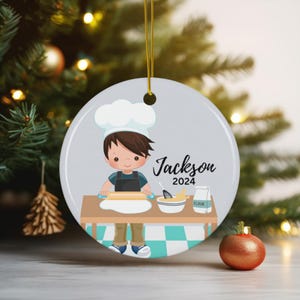 K&ouml;nnte beinhalten: Runder Weihnachtsschmuck mit einem Cartoon-Koch namens Jackson und dem Jahr 2024. Der Koch rollt Teig auf einem Tisch mit einer Sch&uuml;ssel Zutaten und einem Beutel Mehl aus. Der Schmuck hat eine goldene Schnur zum Aufh&auml;ngen.