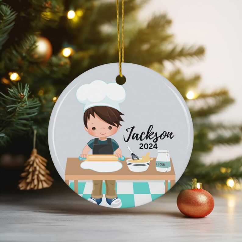 K&ouml;nnte beinhalten: Runder Weihnachtsschmuck mit einem Cartoon-Koch namens Jackson und dem Jahr 2024. Der Koch rollt Teig auf einem Tisch mit einer Sch&uuml;ssel Zutaten und einem Beutel Mehl aus. Der Schmuck hat eine goldene Schnur zum Aufh&auml;ngen.