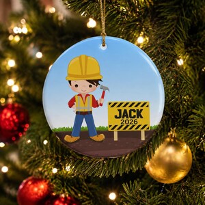 May include: A round ceramic ornament featuring a cartoon construction worker wearing a yellow hard hat, red shirt, and safety vest, holding a hammer. Next to the character is a yellow and black striped sign with the text JACK 2026.