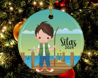May include: A round, white ceramic ornament with a green, blue, and brown illustration of a boy fishing on a dock. The boy is wearing a green vest and blue jeans. The ornament has the name "Silas" and the year "2024" written on it.
