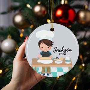 K&ouml;nnte beinhalten: Ein runder Weihnachtsschmuck mit einer Cartoon-Illustration eines jungen Kochs. Der Koch, mit braunen Haaren und einer wei&szlig;en M&uuml;tze, rollt Teig aus. Der Schmuck enth&auml;lt den Namen "Jackson" und das Jahr "2024".