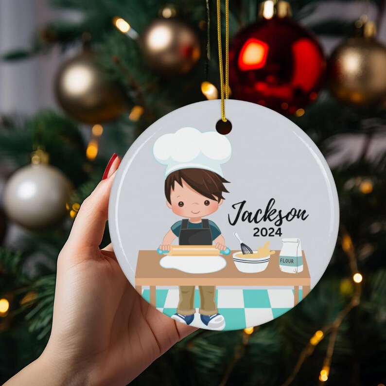 K&ouml;nnte beinhalten: Ein runder Weihnachtsschmuck mit einer Cartoon-Illustration eines jungen Kochs. Der Koch, mit braunen Haaren und einer wei&szlig;en M&uuml;tze, rollt Teig aus. Der Schmuck enth&auml;lt den Namen "Jackson" und das Jahr "2024".