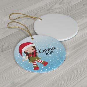 May include: A blue and white Christmas ornament with a cartoon girl wearing a red Santa hat and green elf outfit. The ornament has white snowflakes and the text "Emma 2024".