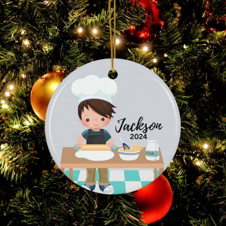 K&ouml;nnte beinhalten: Runder Weihnachtsbaumschmuck aus Keramik mit einem Cartoon-Koch, der Teig ausrollt. Der Schmuck tr&auml;gt den Namen "Jackson" und das Jahr "2024". Er h&auml;ngt an einem goldenen Faden.
