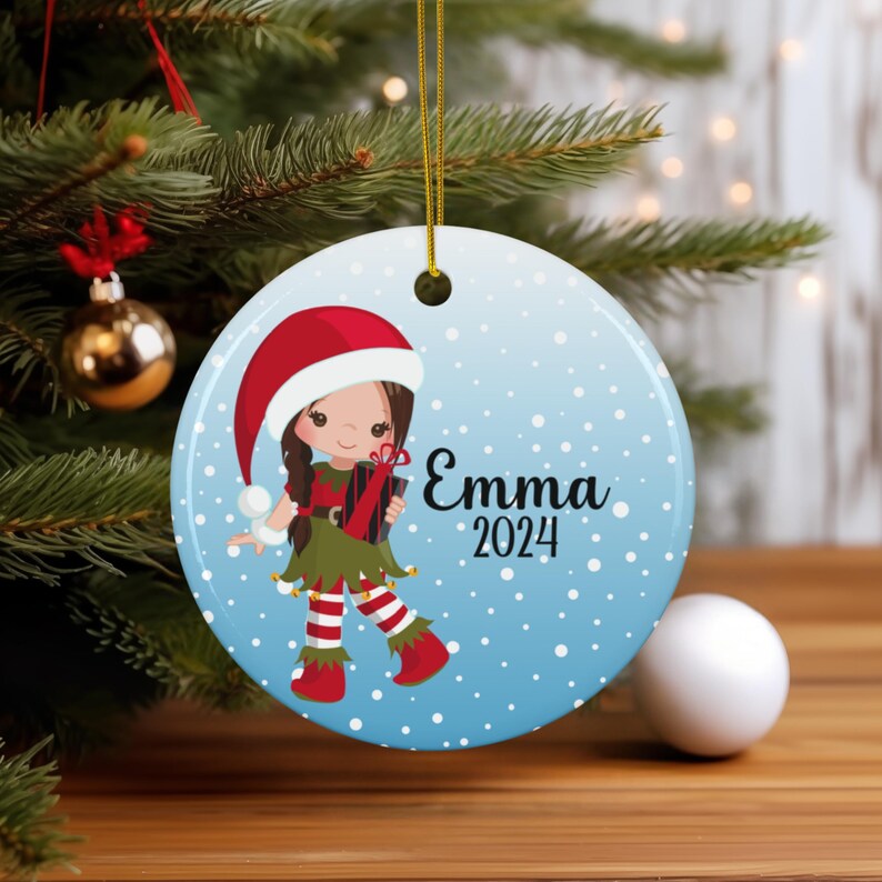 May include: A round blue and white Christmas ornament with a cartoon girl elf holding a gift. The girl has brown hair, a red hat, and green and white striped leggings. The ornament has white snowflakes and the text "Emma 2024".