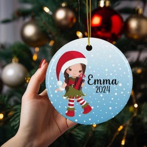 May include: A blue and white Christmas ornament with a cartoon elf girl wearing a red hat and holding a gift. The ornament has white snowflakes and the text "Emma 2024".