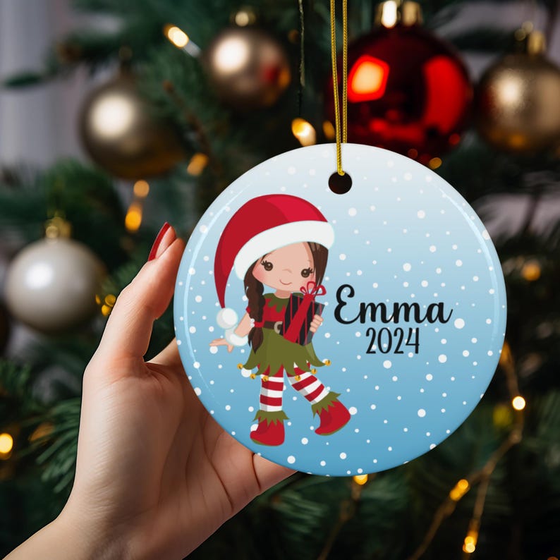 May include: A blue and white Christmas ornament with a cartoon elf girl wearing a red hat and holding a gift. The ornament has white snowflakes and the text "Emma 2024".