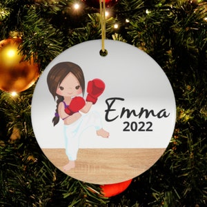 Puede incluir: Adorno navideño redondo y blanco con una niña de dibujos animados en pose de artes marciales, con guantes de boxeo rojos y pantalones blancos. El nombre "Emma" y el año "2022" están impresos en negro. Tiene una cuerda dorada para colgar.