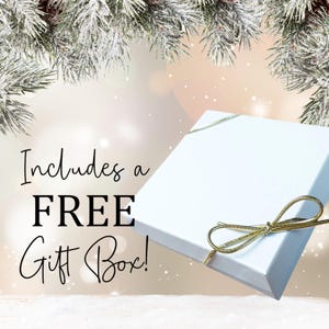 May include: A white gift box with a gold ribbon bow. The box is sitting on a white surface with a snowy pine branch in the background. The text "Includes a FREE Gift Box!" is written in black on a white background.