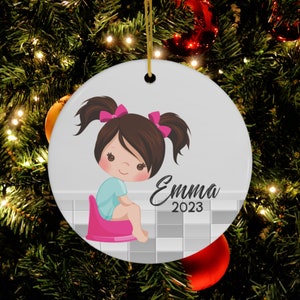 May include: A white ceramic Christmas ornament with a cartoon illustration of a girl with brown hair and pink bows sitting on a pink potty. The ornament has the name "Emma" and the year "2023" written on it.