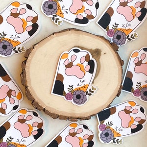 May include: Ten colorful paw print stickers with floral designs. Each sticker features a paw print with black, brown, orange, and white accents. The paw prints are surrounded by stylized flowers in shades of pink, purple, and yellow.