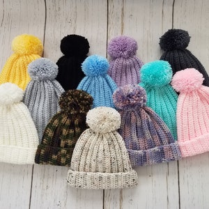 May include: Assortment of 11 crocheted hats with pom-poms in various colors, including white, cream, pink, light blue, gray, purple, black, yellow, and green.