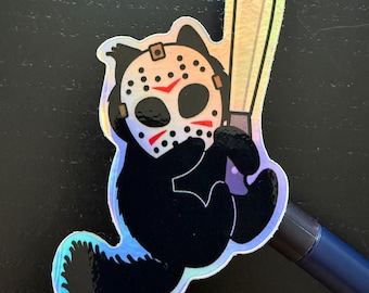 May include: A holographic sticker featuring a black cartoon character wearing a hockey mask with red accents and holding a silver machete. The sticker has a shiny, iridescent finish and is set against a dark background.