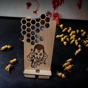 May include: A wooden phone stand featuring a honeycomb design and a portrait of a person with a moustache. The stand is made of light brown wood and has a cutout for a phone.