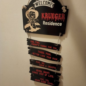 May include: A black wooden sign with a white rope hanger. The sign says "Welcome Krueger Residence". Below the sign are five smaller black wooden signs with red text. The text on the signs reads: "One, Two. Freddy's coming for you. Three, Four. Better lock your door. Five, Six. Grab a crucifix. Seven, Eight. Gonna stay up late. Nine, Ten. Never sleep again..."