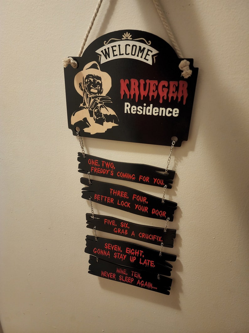 May include: A black wooden sign with a white rope hanger. The sign says "Welcome Krueger Residence". Below the sign are five smaller black wooden signs with red text. The text on the signs reads: "One, Two. Freddy's coming for you. Three, Four. Better lock your door. Five, Six. Grab a crucifix. Seven, Eight. Gonna stay up late. Nine, Ten. Never sleep again..."