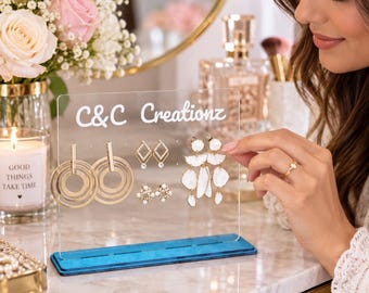 Puede incluir: Un expositor de pendientes de acrílico transparente con base azul, que muestra el texto blanco C&C Creationz. El soporte sostiene tres pares de pendientes: aros dorados, pequeños pendientes de diamante y pendientes colgantes con forma de pétalo blanco.