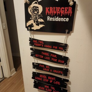 May include: Black wooden sign with the words "Welcome Krueger Residence" in white and red lettering. The sign features a silhouette of Freddy Krueger, a fictional horror character, holding a glove with blades. Below the sign are eight smaller black wooden signs with red lettering, each with a line from the movie "A Nightmare on Elm Street".
