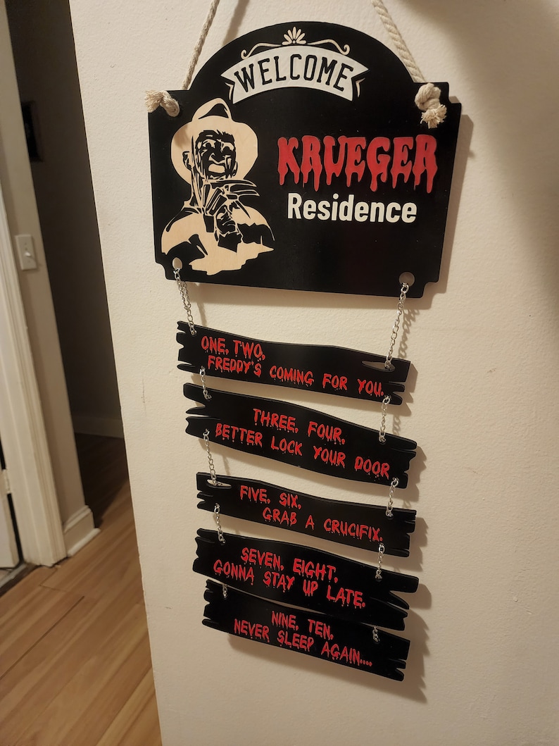 May include: Black wooden sign with the words "Welcome Krueger Residence" in white and red lettering. The sign features a silhouette of Freddy Krueger, a fictional horror character, holding a glove with blades. Below the sign are eight smaller black wooden signs with red lettering, each with a line from the movie "A Nightmare on Elm Street".