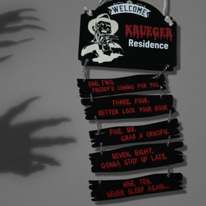 May include: A black wooden sign with a red and white image of Freddy Krueger, the character from the Nightmare on Elm Street horror films. The sign reads "Welcome Krueger Residence" and has a series of smaller signs hanging below with the text "One, two, Freddy's coming for you... Three, four, better lock your door... Five, six, grab a crucifix... Seven, eight, gonna stay up late... Nine, ten, never sleep again..."