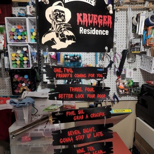 May include: Black wooden sign with a red dripping paint effect that reads "Welcome Krueger Residence".  The sign features a white silhouette of Freddy Krueger with a knife.  Below the sign are five smaller black wooden signs with red text that read: "One, Two, Freddy's Coming For You", "Three, Four, Better Lock Your Door", "Five, Six, Grab A Crucifix", "Seven, Eight, Gonna Stay Up Late", and "Nine, Ten, Never Sleep Again..."