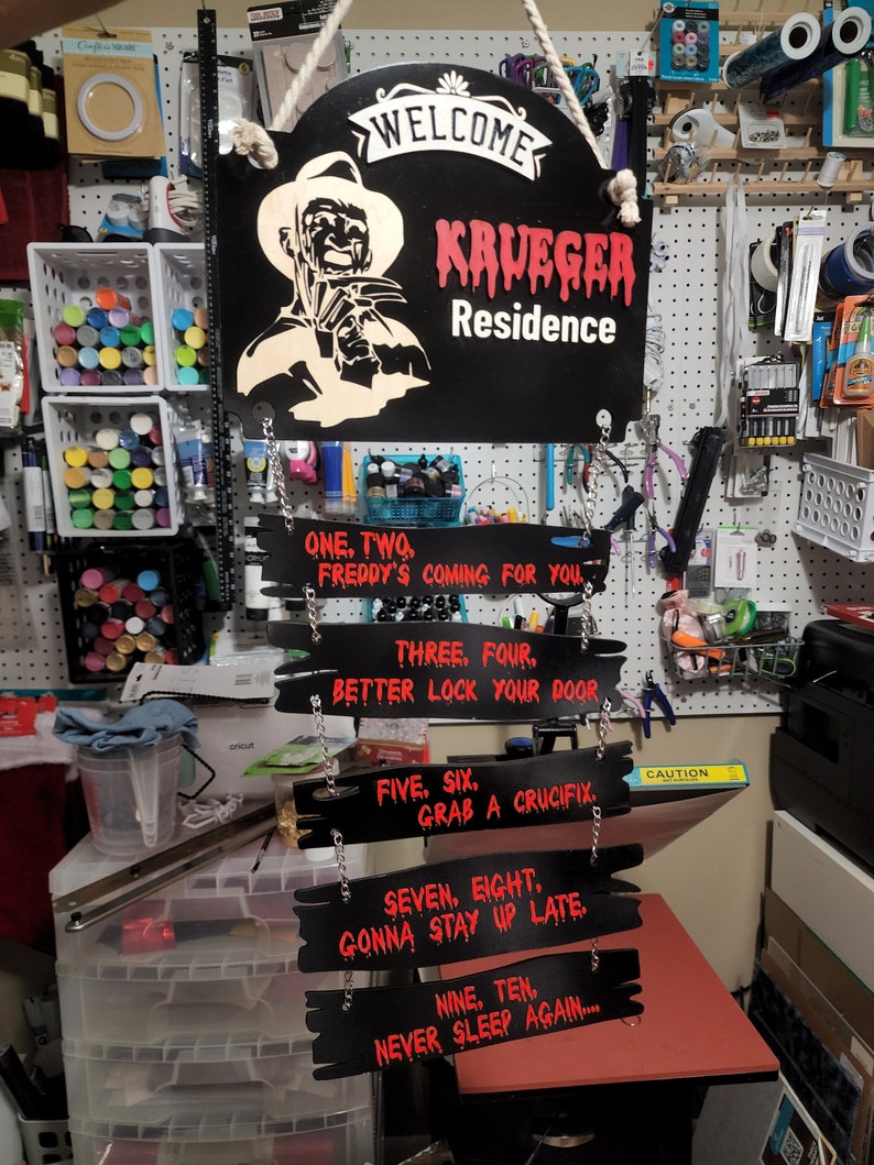 May include: Black wooden sign with a red dripping paint effect that reads "Welcome Krueger Residence".  The sign features a white silhouette of Freddy Krueger with a knife.  Below the sign are five smaller black wooden signs with red text that read: "One, Two, Freddy's Coming For You", "Three, Four, Better Lock Your Door", "Five, Six, Grab A Crucifix", "Seven, Eight, Gonna Stay Up Late", and "Nine, Ten, Never Sleep Again..."