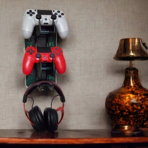 May include: A wall-mounted video game controller holder with two controllers, one red and one white, and a pair of black headphones hanging below. A brown lamp with a gold-coloured shade is on a wooden surface.