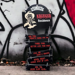 May include: Black and red wooden sign with the text "WELCOME KRUEGER Residence" and a graphic of Freddy Krueger. The sign is hanging from chains and has several smaller signs hanging below it with the text "ONE. TWO. FREDDY'S COMING FOR YOU", "THREE. FOUR. BETTER LOCK YOUR DOOR", "FIVE. SIX. GRAB A CRUCIFIX", "SEVEN. EIGHT. GONNA STAY UP LATE", and "NINE. TEN. NEVER SLEEP AGAIN..."