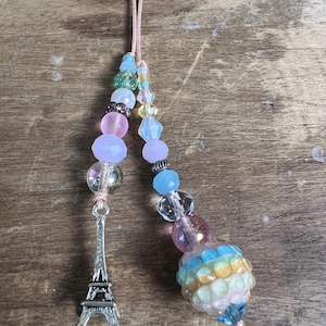 May include: Two beaded charms with pink cords. One charm features a silver Eiffel Tower, the other a colorful, round bead with blue, yellow, and pink accents. Various pastel-colored beads are strung along the cords.