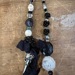 May include: Two beaded pendants with a gothic theme. One features a skull, dark beads, and a silver bird skull charm. The other has black rose beads and a faceted black bead. Both have black ribbon accents and are strung on black cord.