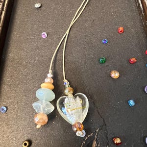 May include: A white heart-shaped glass bead with a silver design and a string of beads with a light green and peach color scheme.