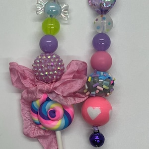 May include: Close-up of two beaded necklaces with colorful beads. One necklace features a pink bow and a lollipop. The other necklace has a heart-shaped bead and a pink bead with white hearts. The beads are in various colors.