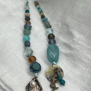 May include: Two beaded bookmarks with a mermaid theme. One bookmark features a seashell charm with a pearl, and the other has a mermaid charm. The beads are in shades of blue, teal, brown, and gold.