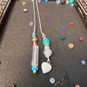 May include: A white heart charm hangs from a beaded necklace with turquoise and gold beads. The necklace is made with a white cord and has a clear crystal pendant.