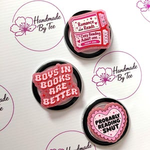 May include: Three circular glittery pins with pink and red accents. One pin is a ticket shape reading Romance Reads. Cozy Reading Admit one. Another reads BOYS IN BOOKS ARE BETTER in red. The third is a heart shape reading PROBABLY READING SMUT.