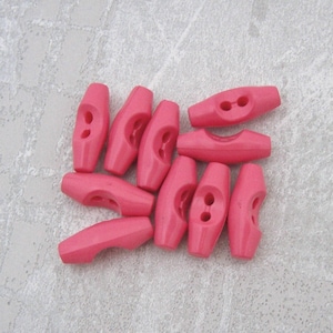 May include: A group of ten pink plastic toggle buttons. The buttons are shaped like a long oval with two holes in the center.