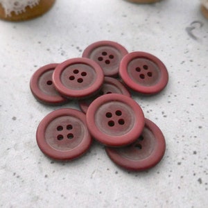 May include: A group of eight maroon buttons with four holes each. The buttons are arranged in a pile on a light gray surface.
