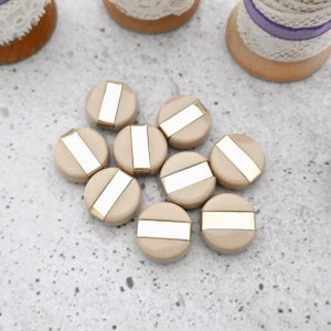 May include: A collection of nine round, beige buttons, each featuring a smooth, glossy finish and a central, horizontal metallic gold-tone bar inlay that spans across the diameter of the button surface.