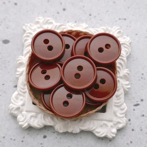 May include: A pile of brown plastic buttons in a white decorative frame. The buttons are all the same size and have two holes.